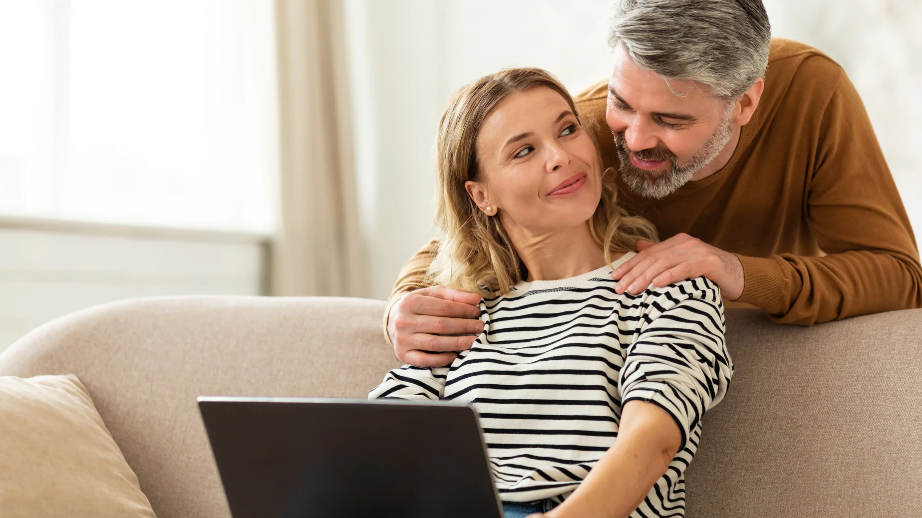 Man embracing wife from behind as she sits on a couch with her laptop
